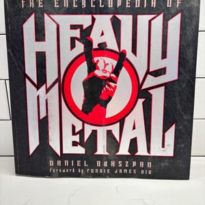 May include: A hardcover book titled The Encyclopedia of Heavy Metal by Daniel Bukszpan with a foreword by Ronnie James Dio. The black cover features large silver lettering, a red circle, and a black hand making the metal horns gesture.