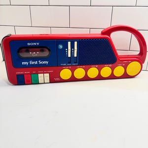 May include: A red and blue My First Sony cassette player featuring a built-in handle, a cassette compartment, volume and master sliders, a speaker grille, and six yellow circular buttons with various animal and drum icons printed on them.