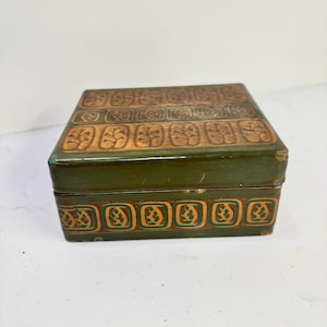 May include: A rectangular box with a dark green finish and a lid featuring a repeating pattern of tan, leaf-like motifs inside square borders. The sides of the box also display a horizontal band of the same green and tan leaf pattern.