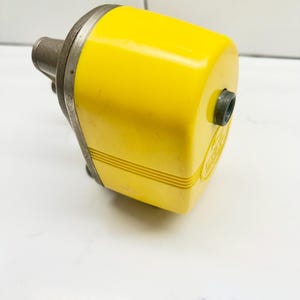 Puede incluir: Un sacapuntas manual de color amarillo brillante con una placa frontal de metal plateado y una pequeña abertura circular en el lateral. La carcasa presenta detalles acanalados horizontales cerca de la base y un emblema circular.