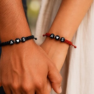 Peut inclure: Deux bracelets en cordon tressé, l'un noir et l'autre rouge, comportant chacun trois perles rondes noires avec des lettres blanches. La perle centrale affiche un cœur blanc, entourée de perles marquées des lettres A et S sur les deux bracelets.