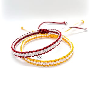 Peut inclure: Deux bracelets en cordon tressé avec fermetures à liens coulissants réglables. Un bracelet est composé de fils rouges et blancs alternés, tandis que l'autre est fait de fils jaunes et blancs. Les lettres ML sont imprimées en noir sur le cordon blanc.