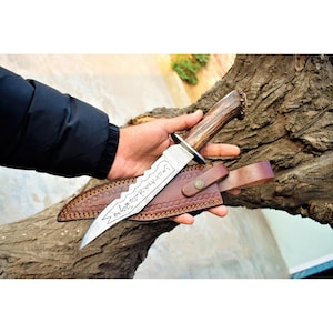 May include: A fixed-blade knife with a textured brown antler handle and a silver-coloured metal guard. The steel blade features a serrated spine and engraved script along the centre. It is accompanied by a brown leather sheath with stitching.