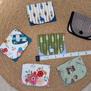 May include: A collection of small, fabric wallets with various patterns, including floral, geometric, and nature-themed designs. Each wallet is closed with a button. A measuring tape is placed across the wallets.