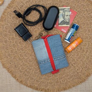 May include: A denim pouch with a red zipper, a black charging cable, a black wall adapter, a black earbud case, a twenty-dollar bill, a red Visa card, a tube of Advil, and an orange lip repair balm. The items are arranged on a round woven mat.
