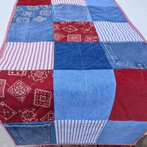 May include: A rectangular table runner featuring a patchwork design of various fabrics including blue denim with pockets, red corduroy, red and white bandana print, and blue and white striped cotton, all finished with a red and white stitched border.