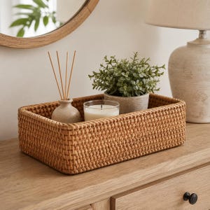 Puede incluir: Una cesta de almacenamiento rectangular de ratán trenzado con un acabado marrón claro natural. Dentro de la cesta hay un pequeño difusor de cerámica beige con varillas de madera, una vela en vaso de cristal y una pequeña planta verde.