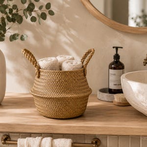 Può includere: Un cesto rotondo in erba marina intrecciata con due manici robusti, riempito con tre asciugamani beige arrotolati ordinatamente. Il cesto presenta un motivo intrecciato testurizzato e poggia su una superficie in legno come soluzione pratica.