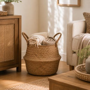 May include: A round, woven seagrass basket with two side handles, featuring a natural tan color and a flexible, collapsible midsection. The basket is filled with several folded, textured beige and cream-colored fabric throws or blankets.