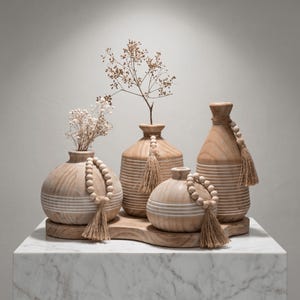 May include: A collection of four wooden vases with a light wood grain finish. Each vase has a unique shape and is adorned with white stripes, wooden bead garlands, and jute tassels. The vases are arranged on a wooden tray, with dried flowers in two of the vases.