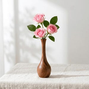 Puede incluir: Un jarrón de madera contiene tres rosas rosas con hojas verdes. El jarrón tiene un cuello delgado y una base redondeada. El arreglo está sobre un mantel texturizado blanquecino, con sombras en la pared blanca.