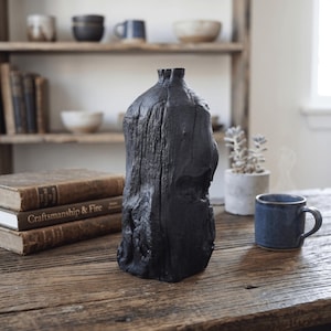 Puede incluir: Un jarrón de madera texturizada de color carbón oscuro con una forma orgánica única. El jarrón tiene un exterior rugoso, similar a la corteza, y una abertura estrecha en la parte superior. Una taza azul y libros están sobre una mesa de madera.