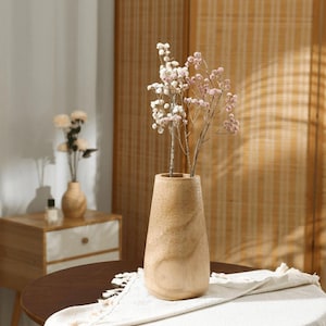 May include: A light brown wooden vase with a wide base and a narrow top, holding delicate pink and white flowers. The vase sits on a dark brown table with a white fringed cloth. A smaller vase is visible in the background.