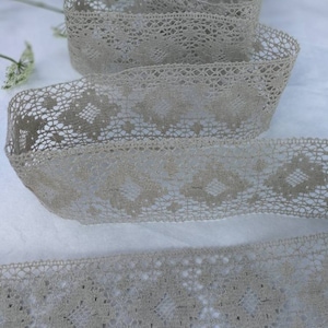 May include: A roll of beige lace ribbon with a delicate, openwork design. The ribbon is tightly rolled, showcasing its intricate patterns and texture. The lace appears soft and flexible, suitable for various craft projects.