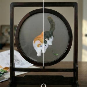 May include: A circular artwork in a dark wooden frame, depicting an embroidered cat. The cat's design is split, with one half orange and white, the other black and white. The artwork is displayed on a wooden surface. Text reads: Dynasty Artisan: The Duality of Color.