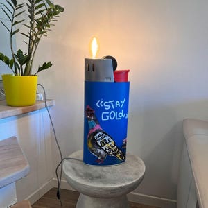 Può includere: Una lampada cilindrica blu a forma di accendino con una parte superiore argentata e una lampadina accesa. Il corpo presenta un'illustrazione colorata di un piccione e la scritta "STAY GOLD" in bianco. Un cavo di alimentazione grigio esce dalla base.