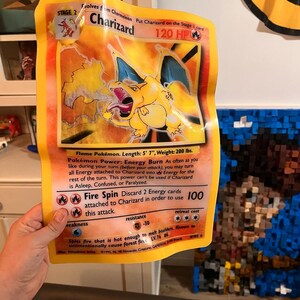 Può includere: Una grande carta Pokémon Charizard gialla. La carta presenta un'illustrazione infuocata arancione e gialla di Charizard, con testo che indica i suoi PS, attacchi e abilità. La carta è un'evoluzione di livello 2, con una lunghezza di 170 cm e un peso di 91 kg.