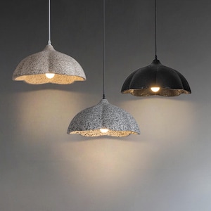 May include: Three pendant lights with textured, dome-shaped shades. The shades are in shades of grey and black, with a scalloped edge. Each light has a single bulb illuminating the interior. The lights hang from black or white cords.