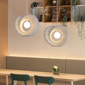 May include: Two circular pendant lights with a speckled grey stone-like texture. Each fixture features a central circular opening containing a single glowing white spherical light bulb, suspended by a thin white cord from the ceiling above.