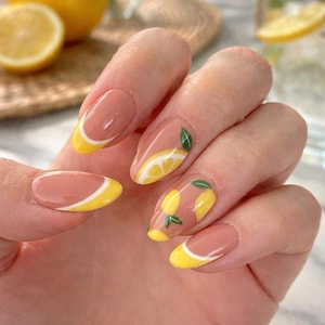 May include: Close-up of a hand with almond-shaped nails. The nails feature a nude base with yellow and white accents. Some nails have a lemon slice design with green leaves, while others have a yellow and white curved design.