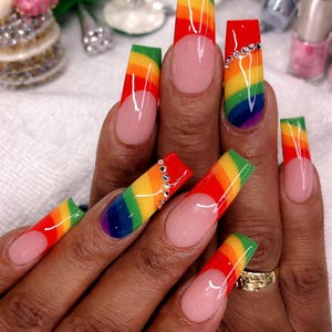 May include: Long, square-shaped acrylic nails with a rainbow gradient design. Some nails feature a clear base with a rainbow stripe, while others have a red tip and are adorned with rhinestones. The nails are a vibrant and colorful fashion statement.