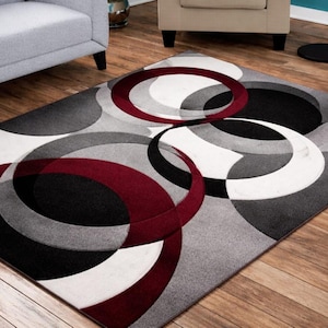 May include: A rectangular rug with a contemporary design. It features overlapping circles in grey, black, white, and burgundy. The rug is placed on a wooden floor, with furniture visible in the background.