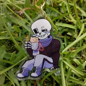May include: A clear acrylic keychain featuring a cartoon skeleton character with a white skull, wearing a dark purple sweater and light purple pants, holding a small mug with a cat face design, attached to a gold-colored metal star-shaped clasp.