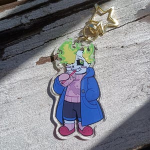May include: A clear acrylic charm featuring a cartoon skeleton character with bright green hair, wearing a blue coat over a pink sweater and dark shorts, holding a pink drink. The charm includes a gold-colored star-shaped metal clasp at the top.