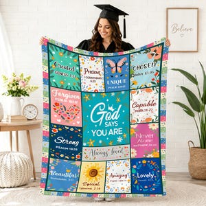 May include: A colorful, patchwork-style blanket with inspirational text and floral designs. The blanket features phrases like "Created," "Precious," and "God Says You Are." The design includes a butterfly, a sunflower, and various floral elements. The blanket is held up by a person.