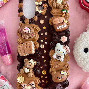 K&ouml;nnte beinhalten: Transparente Handyh&uuml;lle mit braunen und beigen geformten Elementen. Die H&uuml;lle zeigt eine Vielzahl von Miniaturfiguren, darunter Teddyb&auml;ren, ein H&auml;schen, ein Affe und eine Hello Kitty-Figur. Das Wort "COCO" ist sichtbar.