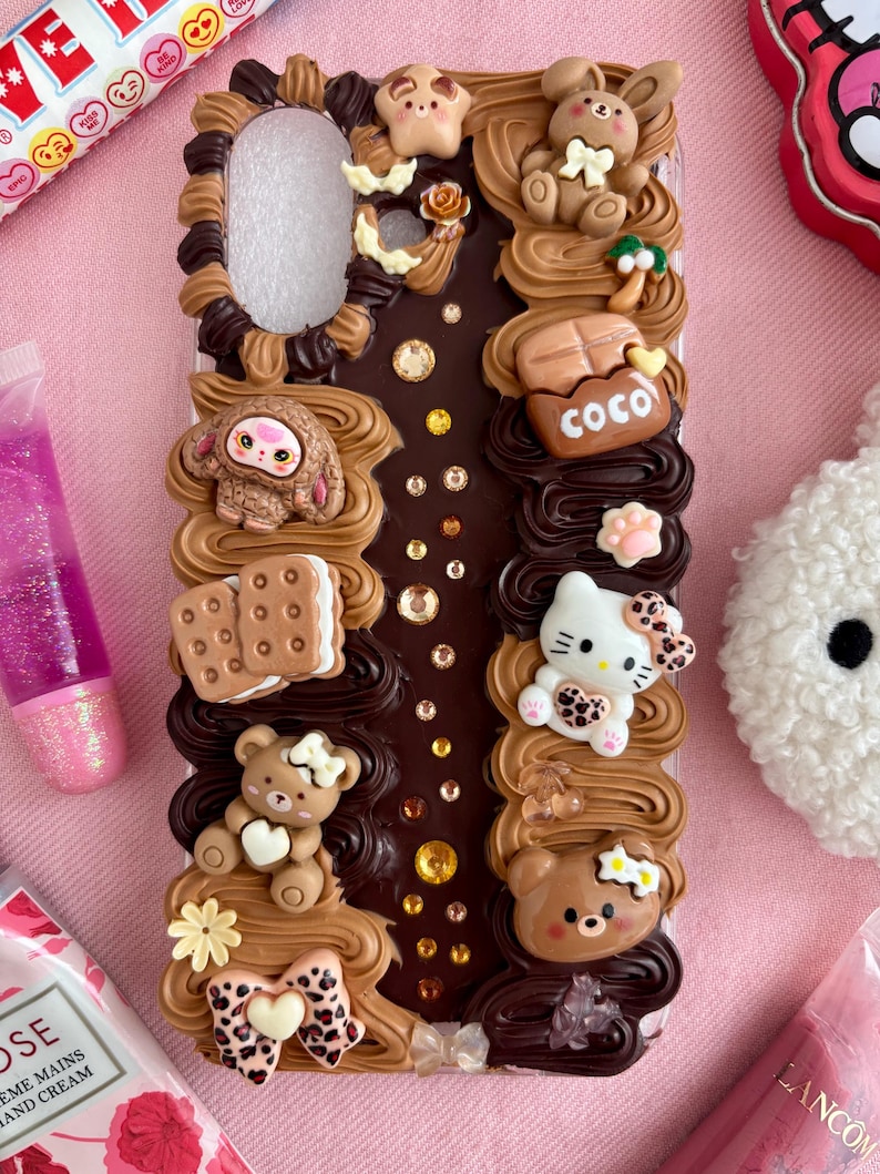 K&ouml;nnte beinhalten: Transparente Handyh&uuml;lle mit braunen und beigen geformten Elementen. Die H&uuml;lle zeigt eine Vielzahl von Miniaturfiguren, darunter Teddyb&auml;ren, ein H&auml;schen, ein Affe und eine Hello Kitty-Figur. Das Wort "COCO" ist sichtbar.