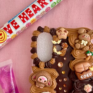 K&ouml;nnte beinhalten: Eine dekorative Handyh&uuml;lle mit einem Schokoladen-Design, mit einem Hasen, einem Schaf und anderen niedlichen Elementen. Eine Tube Love Hearts-Bonbons und eine Tube rosa Lipgloss sind ebenfalls im Bild.