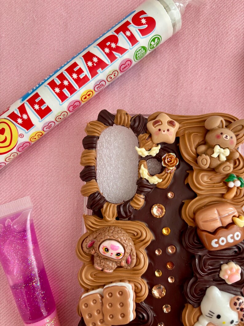 K&ouml;nnte beinhalten: Eine dekorative Handyh&uuml;lle mit einem Schokoladen-Design, mit einem Hasen, einem Schaf und anderen niedlichen Elementen. Eine Tube Love Hearts-Bonbons und eine Tube rosa Lipgloss sind ebenfalls im Bild.