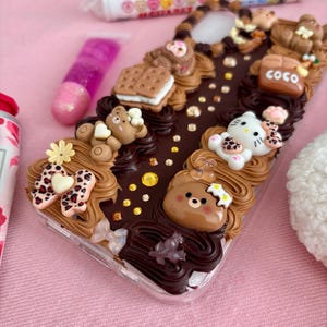K&ouml;nnte beinhalten: Eine transparente Handyh&uuml;lle, verziert mit braunem Zuckerguss, Miniatur-Teddyb&auml;ren, einer Hello Kitty-Figur und einem Keks. Die H&uuml;lle hat auch eine Schleife mit Leopardenmuster und das Wort "COCO".