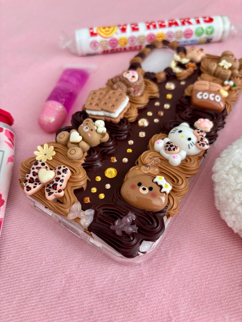 K&ouml;nnte beinhalten: Eine transparente Handyh&uuml;lle, verziert mit braunem Zuckerguss, Miniatur-Teddyb&auml;ren, einer Hello Kitty-Figur und einem Keks. Die H&uuml;lle hat auch eine Schleife mit Leopardenmuster und das Wort "COCO".