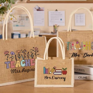 Può includere: Tre borse di tela di iuta con manici bianchi. Le borse presentano la parola "TEACHER" in testo colorato con grafica floreale e scolastica. I nomi "Mrs. Regina", "Mrs. Barbie" e "Mrs. Darcey" sono stampati sotto.