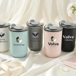 Könnte beinhalten: Fünf zylindrische Becher mit Deckeln in den matten Farben Weiß, Hellblau, Dunkelgrau, Zartrosa und Schwarz. Jeder Becher zeigt ein individuelles Logo, darunter Veyra Collective und Volve Collective, sowie einen grauen Schiebedeckel-Mechanismus.