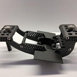 Puede incluir: Un soporte de cámara impreso en 3D de color negro con una superficie texturizada que presenta numerosos recortes circulares, dos plataformas de montaje planas con perillas de metal plateado y una carcasa central rectangular, ensamblado con tornillos plateados.