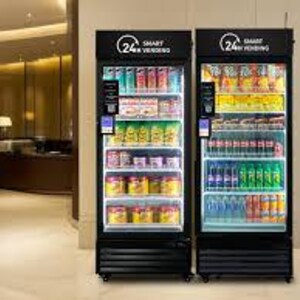May include: Two black smart vending machines with glass doors, filled with various beverages and snacks. The machines have the text "24h Smart Vending" at the top. The interior is brightly lit, showcasing the products on multiple shelves.