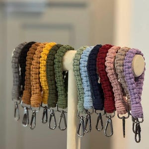 Puede incluir: Una colección de correas de muñeca de macramé en varios colores, incluyendo gris, negro, marrón, amarillo, verde, azul, marino, rojo, rosa, beige y morado. Cada correa está unida a un cierre metálico plateado.