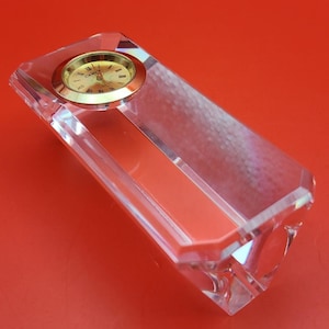 May include: A clear crystal desk clock with a gold-coloured clock face. The clock has a rectangular shape with a beveled edge and sits on a red surface. The clock face has Roman numerals and the brand name "CAMLY".