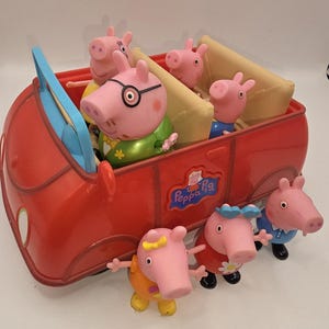 Puede incluir: Un coche de juguete de plástico rojo con el logotipo de Peppa Pig en el lateral, que contiene cuatro figuras de cerditos en su interior. Tres figuras de cerditos adicionales están de pie frente al vehículo. Las figuras son rosas con ropa variada.