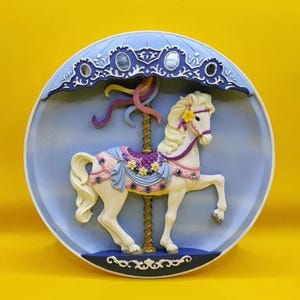 May include: A carousel horse sculpture in a circular frame. The horse is white with a yellow flower in its mane, adorned with a purple saddle and pink and blue accents. The frame is light blue with a dark blue decorative top.