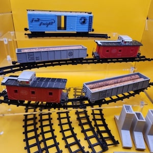 May include: A collection of toy train cars and tracks. The set includes a blue freight car with "Fast Freight" text, red cabooses, and gray freight cars. The tracks and accessories are black and gray.