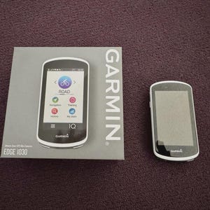 May include: A Garmin Edge 1030 GPS bike computer and its box. The computer is black and white with a touchscreen display showing navigation and training options. The box is gray with the Garmin logo and the Edge 1030 model name.