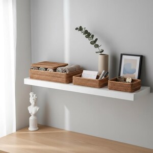 May include: A collection of woven rattan storage baskets in various sizes, displayed on a white shelf. The baskets contain rolled towels, stationery, and small decorative items. A framed artwork and a vase with greenery are also on the shelf.
