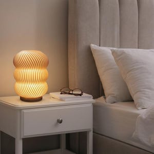 May include: A bedside lamp with a warm, inviting light. The lamp features a distinctive, layered design with vertical grooves, placed on a white bedside table. A pair of spectacles sits on a stack of books.