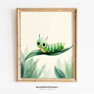 May include: A framed watercolor art print featuring a cute green caterpillar with large black eyes and a small smile resting on a long green leaf, set against a pale cream background with soft green foliage at the bottom of the composition.
