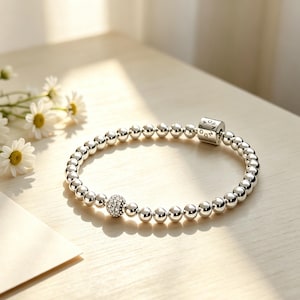 Puede incluir: Una pulsera de cuentas plateadas que presenta un dije esférico incrustado con piedras transparentes y un cierre cilíndrico de metal grabado con el texto PANDORA. La pulsera está compuesta por esferas metálicas pulidas y reflectantes.