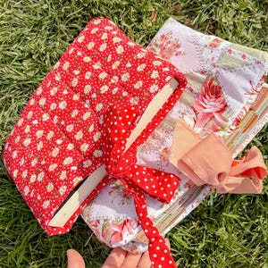 May include: Two fabric book covers with floral and polka dot patterns. One cover is red with white daisies, the other is patchwork with pink roses. A red polka dot ribbon ties the red cover closed. The covers are on a bed of green grass.